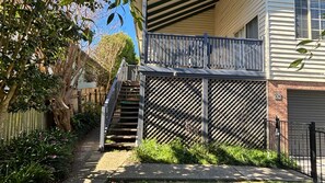 Un immeuble résidentiel avec un escalier en bois menant à un balcon, une porte de garage et une cour clÎturée agrémentée de verdure.