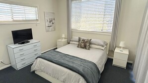 A bedroom with a bed, a TV on a dresser, and a window with blinds. - Susie's Beach House - South West Rocks (SOUTH WEST ROCKS)