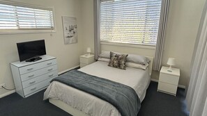 Ein Schlafzimmer mit Bett, Fernseher auf einer Kommode, Nachttisch und Fenster mit Jalousien.