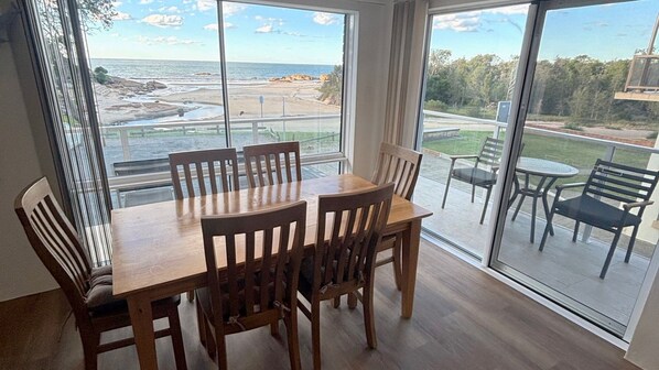 Un comedor con mesa y sillas, ventanas amplias con vista a la playa y un balcón con mobiliario exterior.
