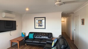 Una sala de estar con un sofá de cuero negro, una mesa de centro de madera, un televisor de pantalla plana montado en la pared y un ventilador de techo.