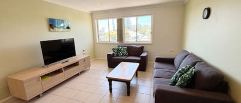 Een woonkamer met een flatscreen televisie, een houten salontafel, twee banken en een raam met uitzicht op bomen en gebouwen.
