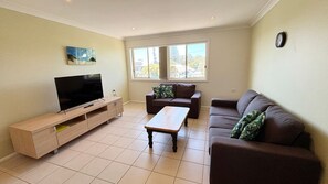 Una sala de estar con una televisión de pantalla plana, una mesa de centro de madera, dos sofás y una ventana que ofrece vistas a árboles y edificios.