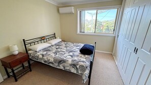 A single bed with a floral bedspread, a bedside table with a lamp, and a window with a view of greenery.