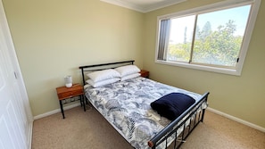 A single bed with a patterned bedspread, a bedside table with a lamp, and a window with a view of trees. - Village Centre 3 - South West Rocks (SOUTH WEST ROCKS)