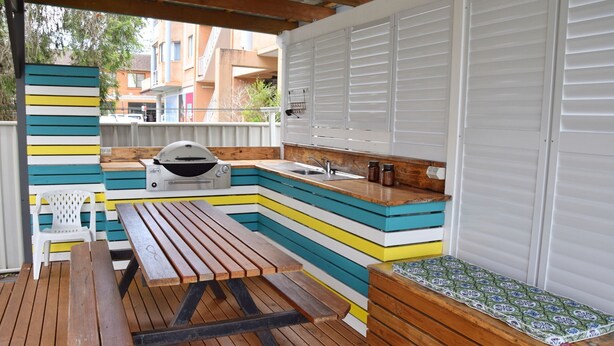 Una cocina exterior compacta con una parrilla, pileta y mesa de madera, rodeada por una terraza colorida.