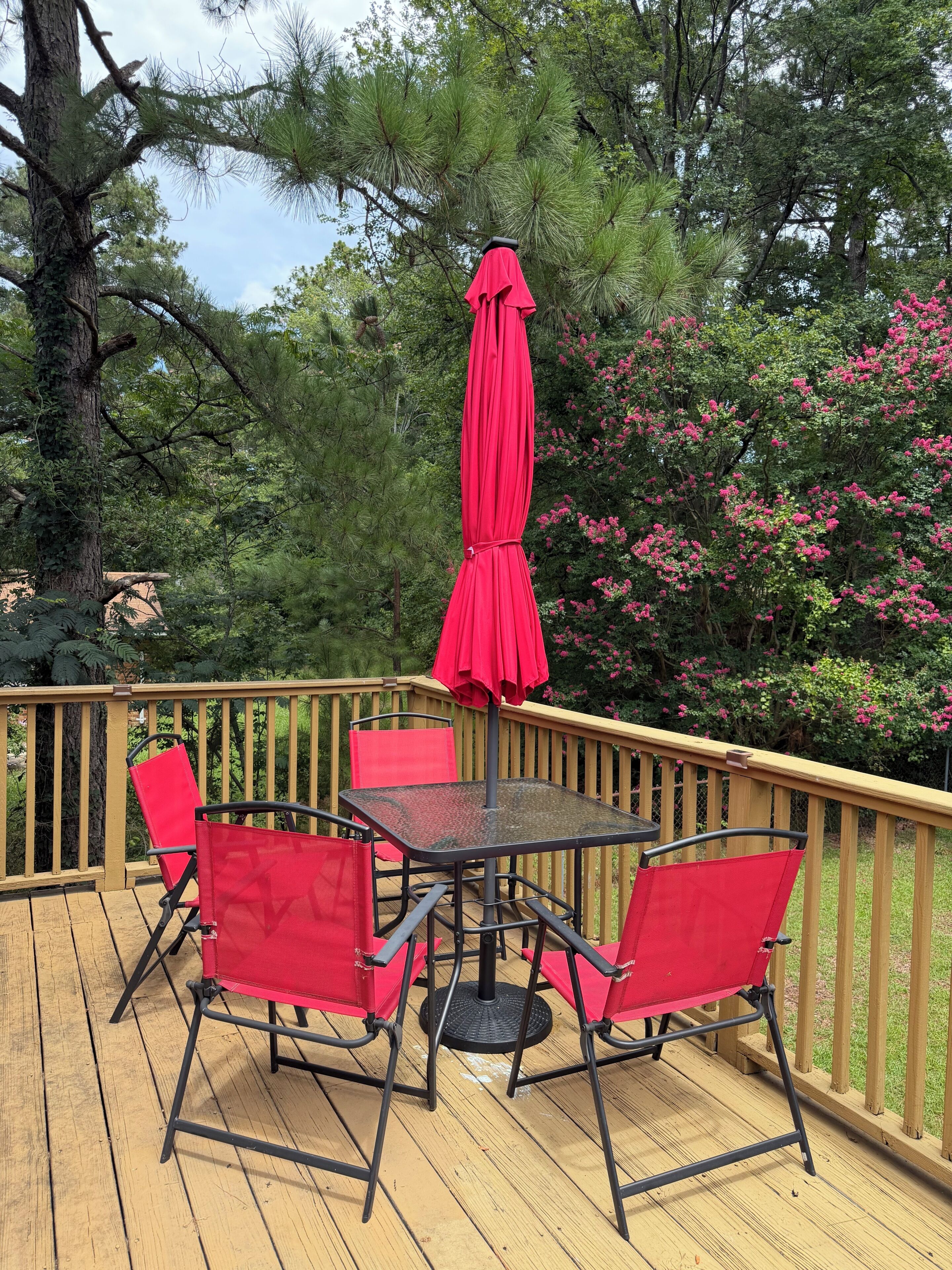 Una terraza con una mesa de vidrio y cuatro sillas rojas, rodeada por una barandilla de madera y abundante vegetación.