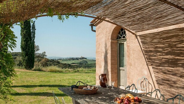 Un coin repas extérieur rustique, avec une table dressée pour deux personnes, un panier de pain, une carafe et un saladier rempli de fruits.