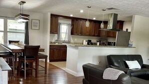 Un salon moderne avec un canapé en cuir foncé, un coin repas avec des chaises en bois et une cuisine dotée de meubles en bois et d’un plan de travail en granit.