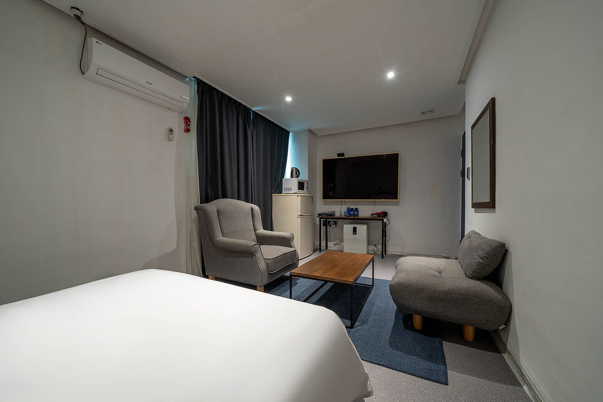 A hotel room with a bed, two armchairs, a small wooden table, a desk with a computer and a microwave, and a TV on the wall.