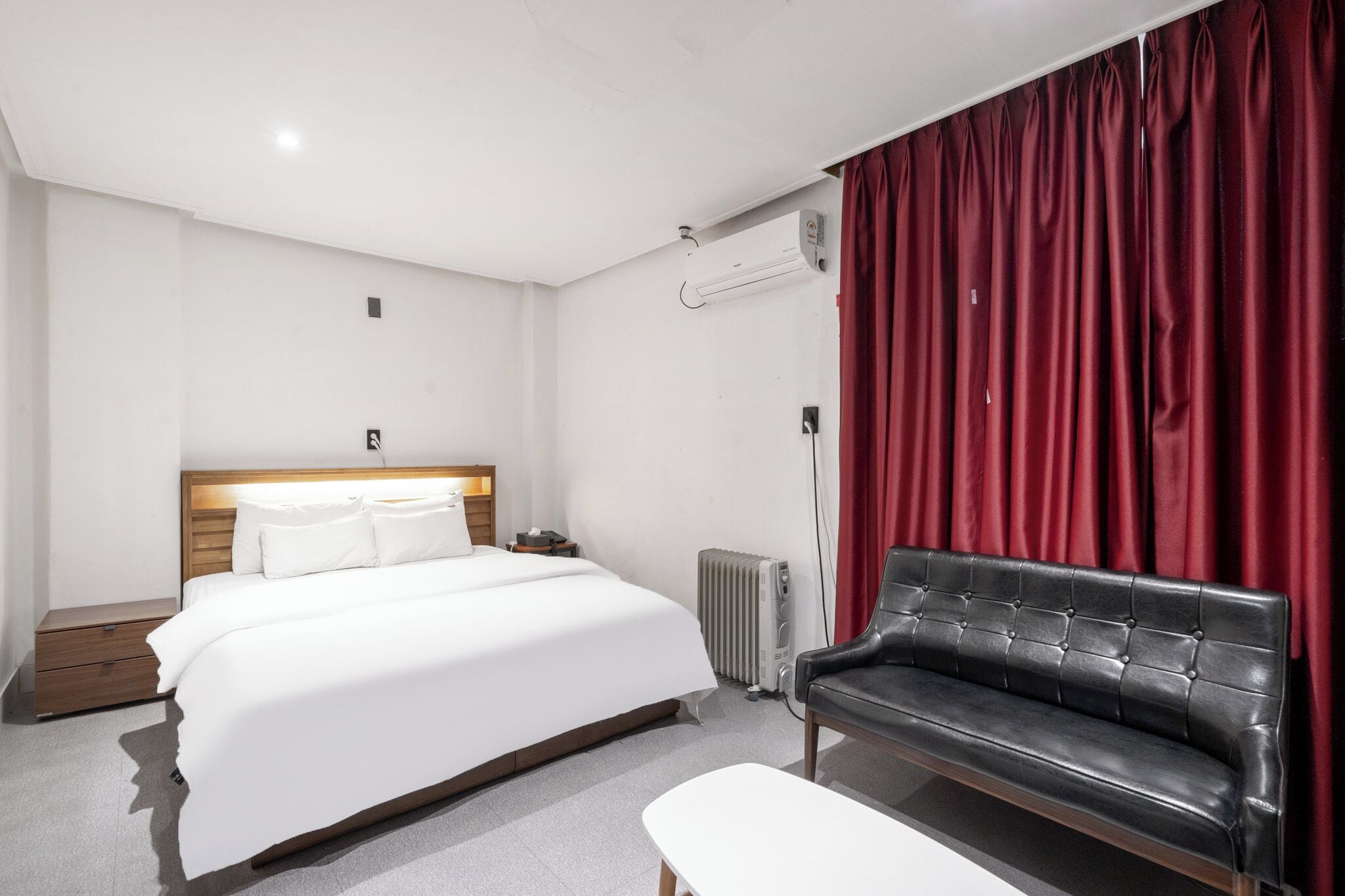 Una habitación de hotel con cama, mesita de noche, sofá de cuero negro, cortina roja y radiador.