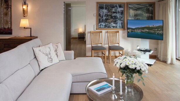 A modern living room with a white sofa, two wooden chairs, a coffee table with a book and candles, a TV, and artwork on the walls. - Chesa Piz Padella - St. Moritz by Interhome (St. Moritz)