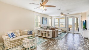 A spacious living room with a beige checkered sofa, a coffee table, and a TV on a stand. There are two ceiling fans, a large window with blinds, and a view of the outside balcony.