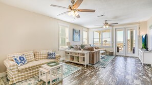 A spacious living room with a beige checkered sofa, a coffee table, and a TV on a stand. There are two ceiling fans, a large window with blinds, and a view of the outside balcony.