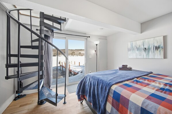 A bedroom with a bed, a blue blanket, a checkered bedspread, a painting on the wall, and a staircase leading to a balcony. - NEW Lakefront Escape Private Dock & Lake Views (Alton)