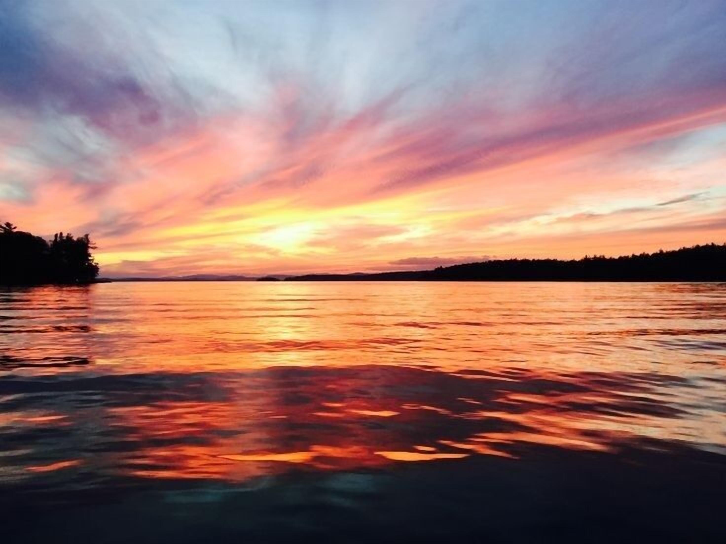 Ein Sonnenuntergang über ruhigem Wasser, im Horizont die Silhouette von Bäumen.