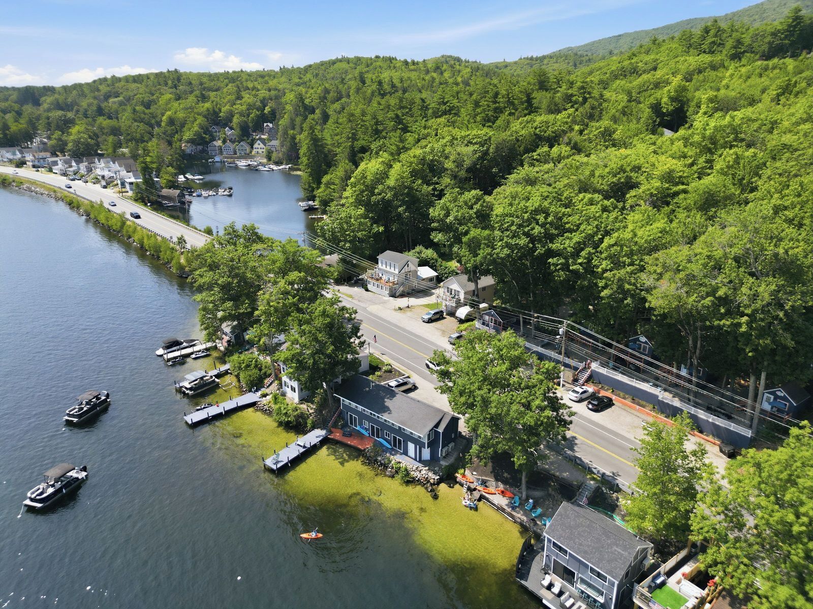 A lakeside property with docked boats, a road, and a forested hill.