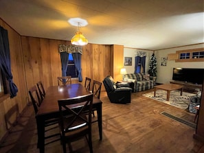 Un salon chaleureux avec des murs en bois, une table à manger, des chaises, un canapé, un téléviseur et un sapin de Noël.