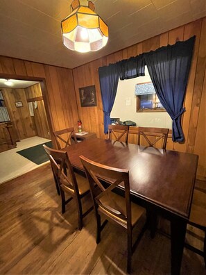 Une salle à manger aux murs lambrissés, avec une table ronde et des chaises, un luminaire suspendu et des rideaux bleus.