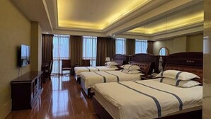 Quarto de hotel com três camas, televisão de tela plana, cómoda de madeira e janelas grandes com cortinas.