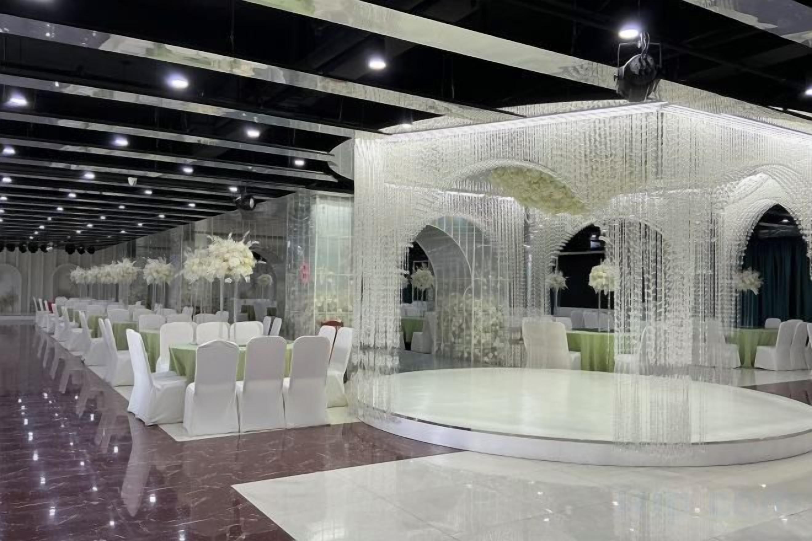 Un salón de banquetes con un escenario central, sillas blancas y centros de mesa florales.