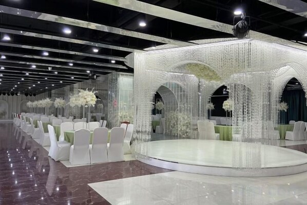 Un salón de banquetes con un escenario central, sillas blancas y centros de mesa florales.
