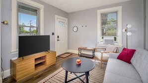 Una sala de estar moderna con un televisor de pantalla plana sobre un soporte de madera, un sofá gris con un cojín rojo, una pequeña mesa redonda y una ventana con persianas.