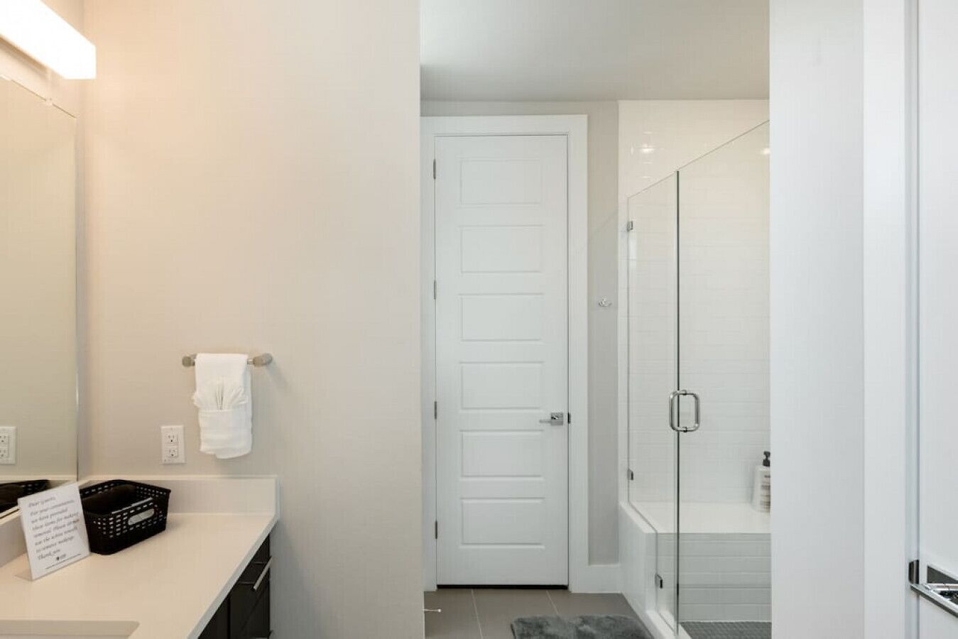 A modern bathroom with a white door, a glass shower enclosure, and a white countertop with a basket and a towel.
