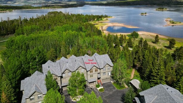 A large, multi-story building with a grey roof and multiple windows, surrounded by dense green forest and a lake. - Prospector Point E 4br 3.5ba 4 Bedroom Townhouse by RedAwning (Frisco)