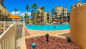 Zona de piscina con una piscina azul cristalina, rodeada de palmeras y edificios con balcones.
