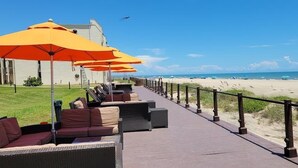 Un paseo marítimo frente a la playa con asientos al aire libre y sombrillas, una barandilla y vistas a la playa y el océano.