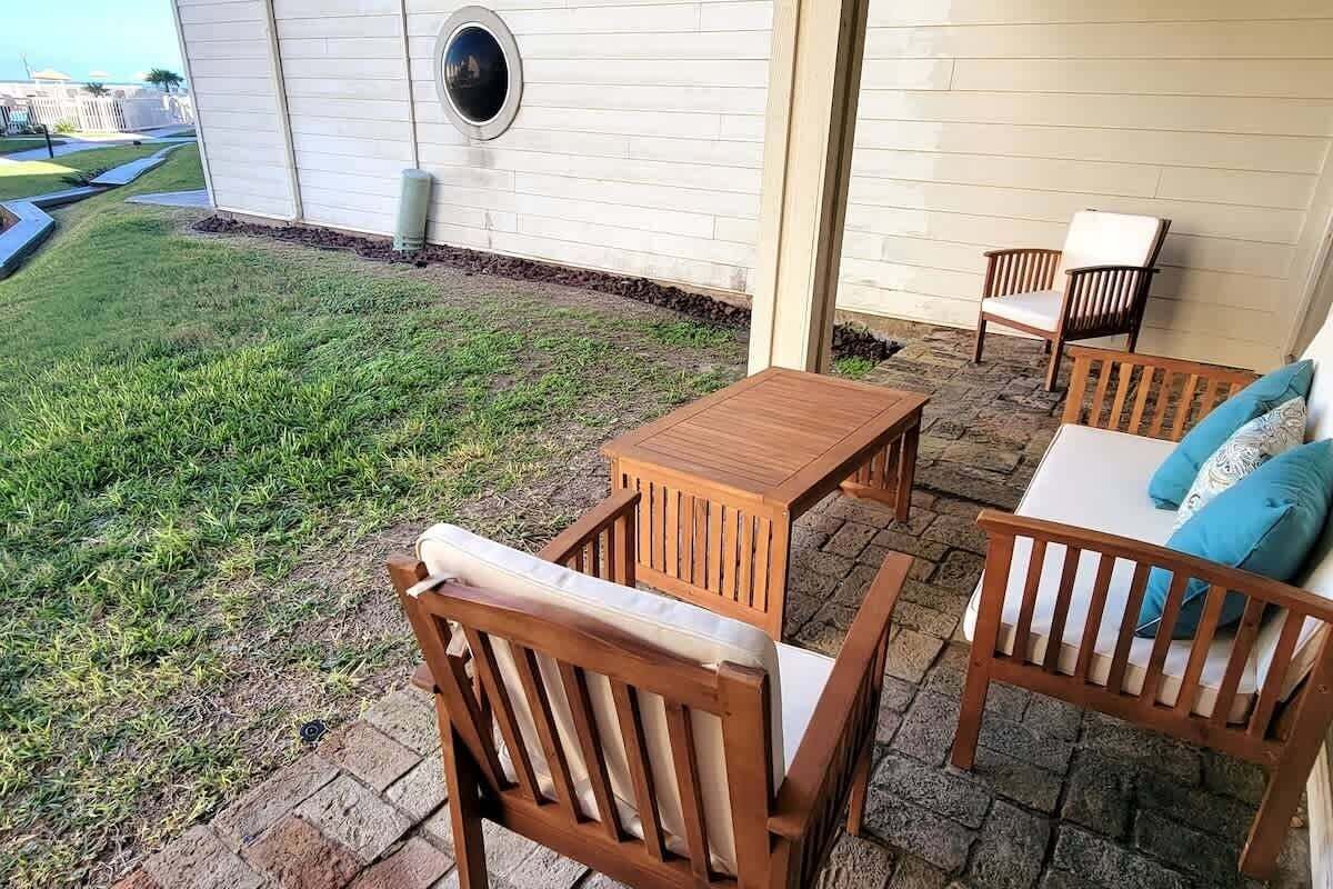 Une terrasse dotée de mobilier en bois, comprenant une table et des chaises, et un mur blanc orné d’une fenêtre circulaire.