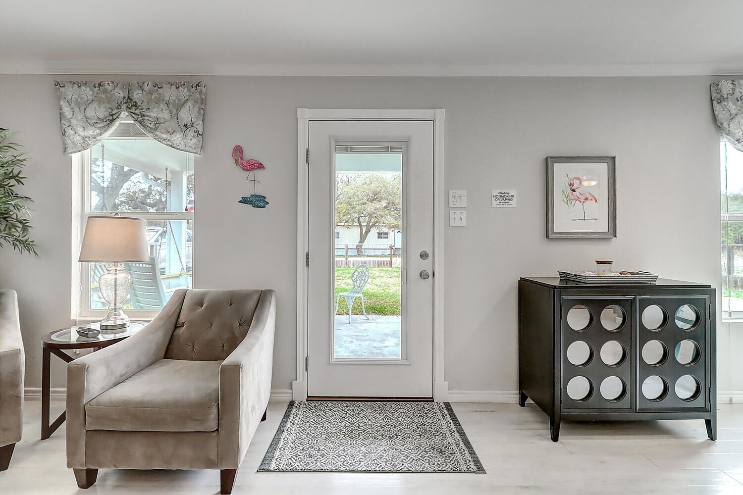 A living room with a beige armchair, a side table with a lamp, a dark cabinet with circular mirrors, and a door with a glass panel.