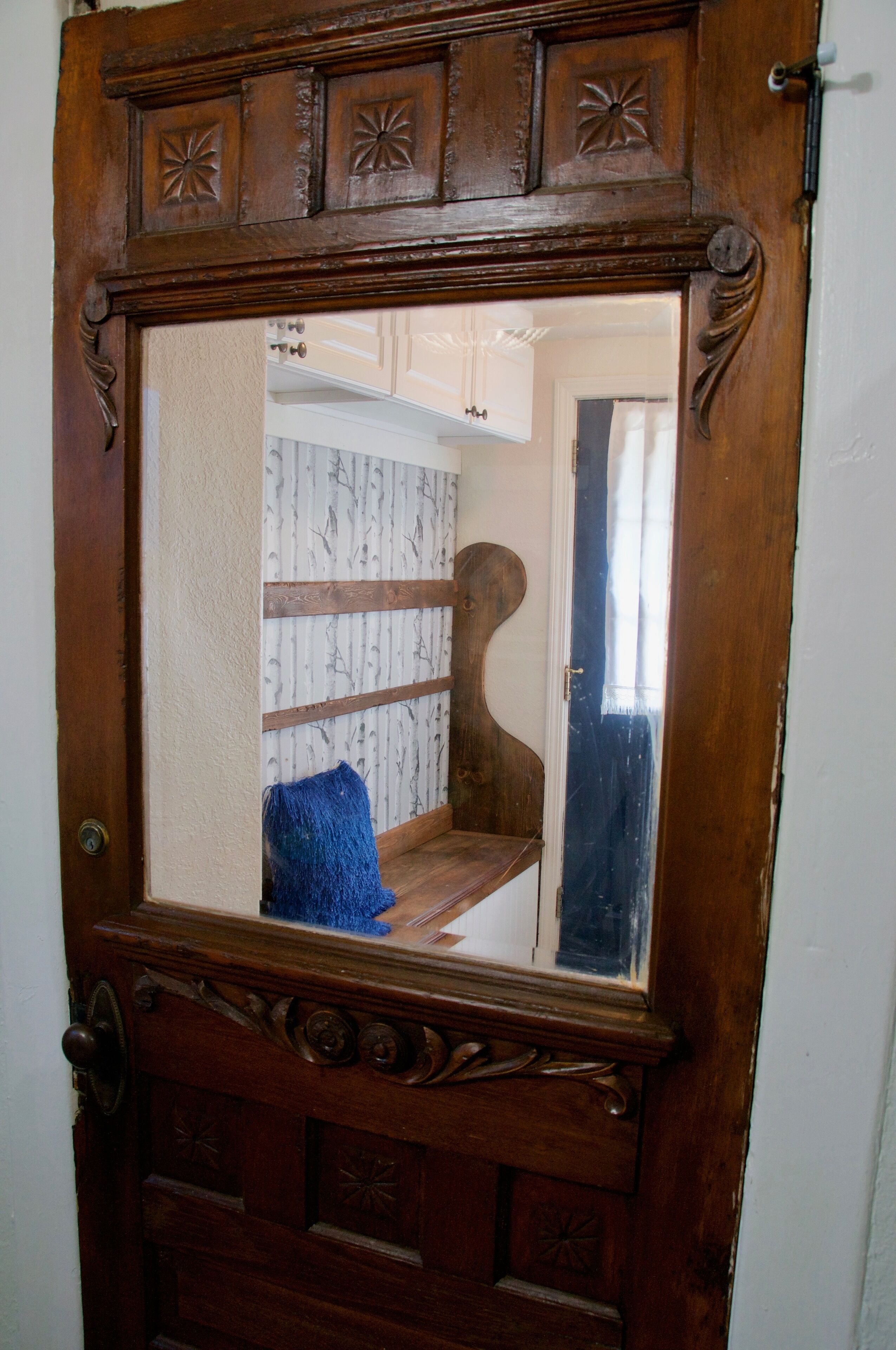 A wooden-framed mirror with a view of a room featuring a patterned wallpaper and a wooden bench.