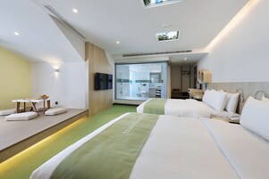Una habitación de hotel moderna con dos camas, una mesita con un frutero, y vistas al baño y a la cocina a través de una puerta de vidrio.