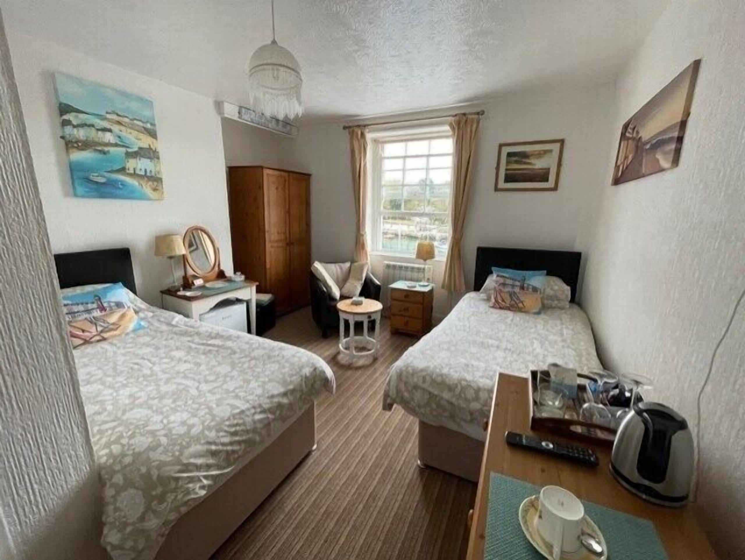 A hotel room with two beds, a desk, a chair, a small table, a window with curtains, and artwork on the walls.