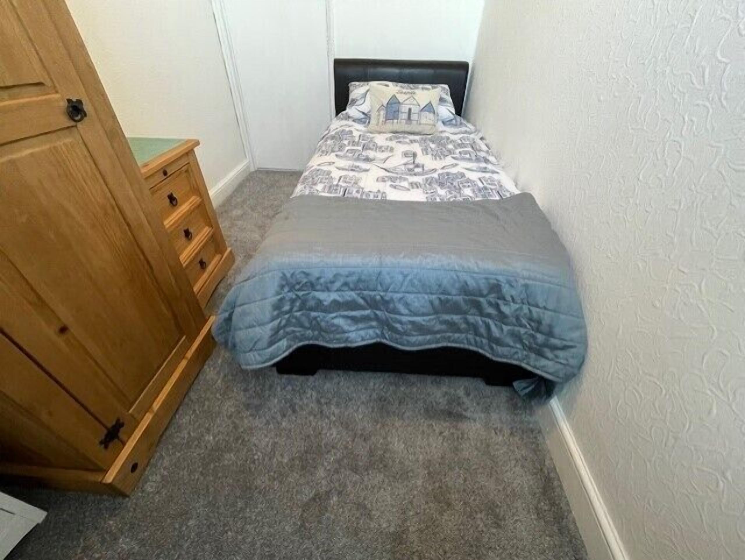 A small bedroom with a single bed, a wooden wardrobe with three drawers, and a plain carpeted floor.