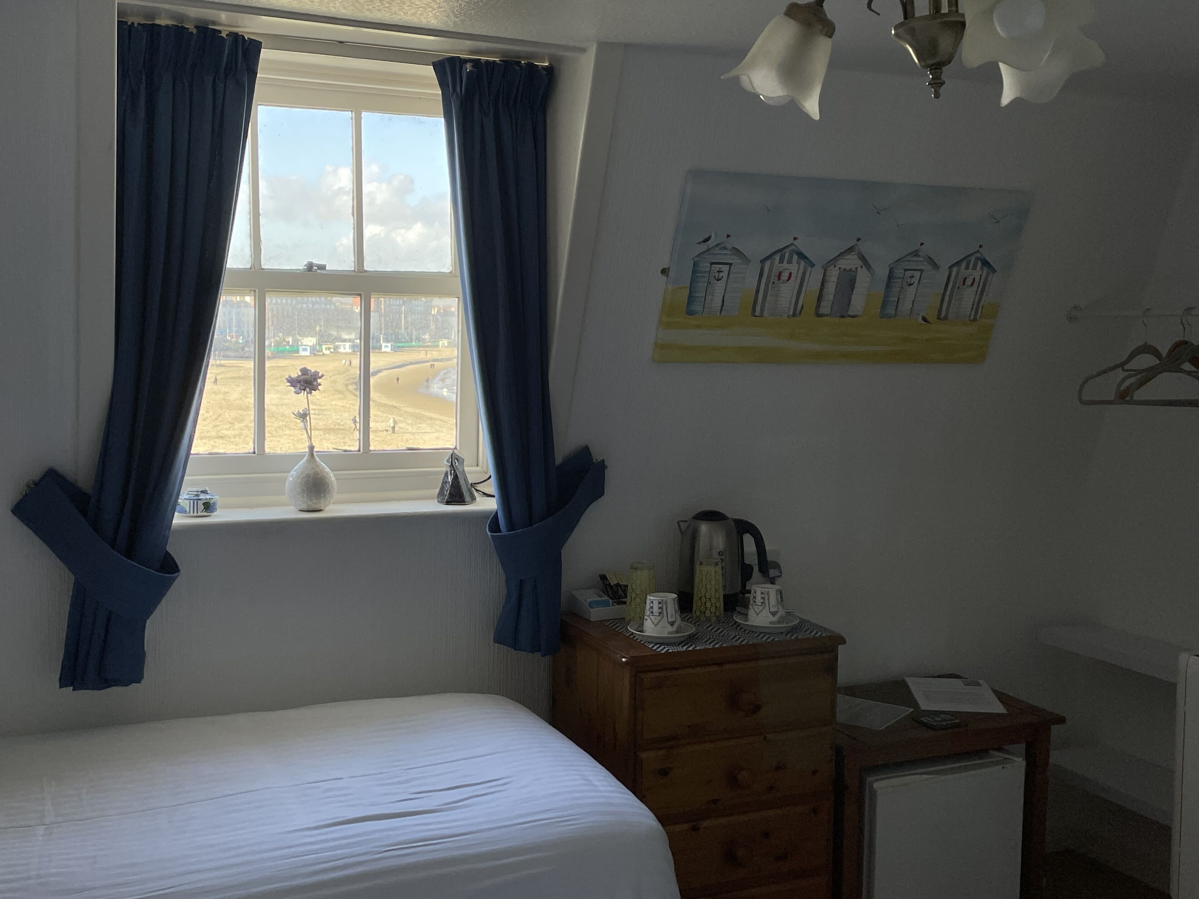 Een slaapkamer met een bed, een kaptafel, een bureau, een radiator, een raam met gordijnen, een schilderij van strandhutten en uitzicht op het strand.