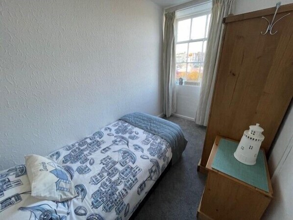 Une petite chambre avec un lit simple, une couvre-lit à motifs, une armoire en bois et une petite table sur laquelle se trouve un objet blanc.