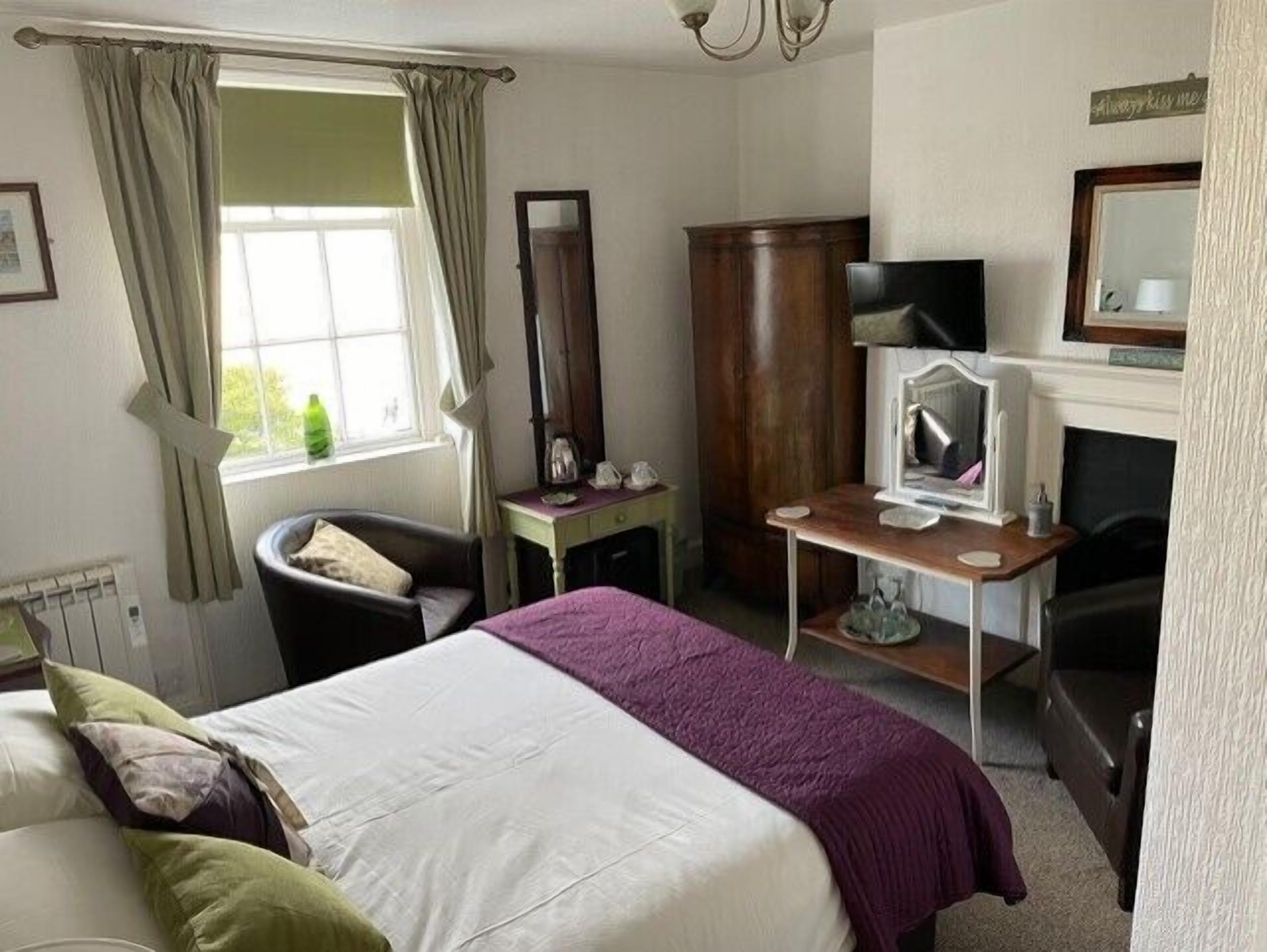 A bedroom with a bed, a chair, a desk, a mirror, and a fireplace.