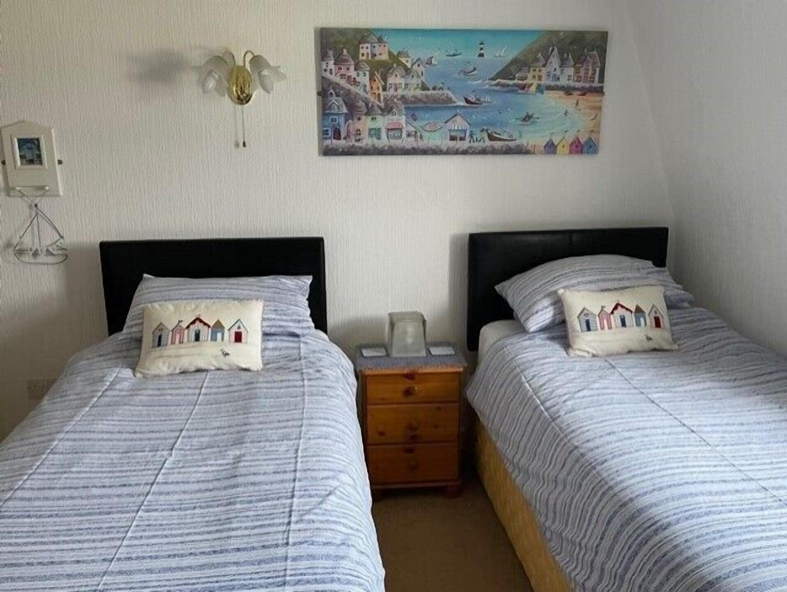 A room with two beds, each with a pillow featuring a coastal scene. A painting of a coastal village hangs on the wall above the beds.