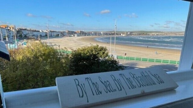 Vista d'una platja des d'una finestra amb un cartell que diu 'By The Sea By The Beach'.