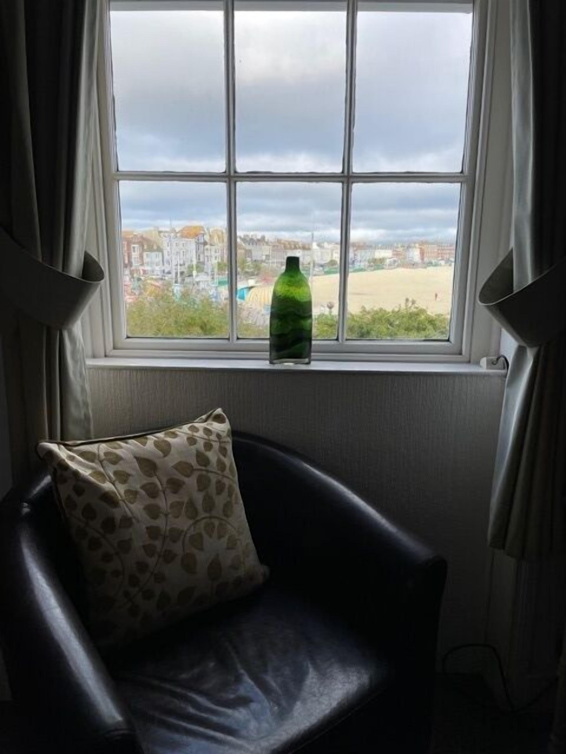 A chair with a patterned pillow in front of a window with a bottle on the sill.