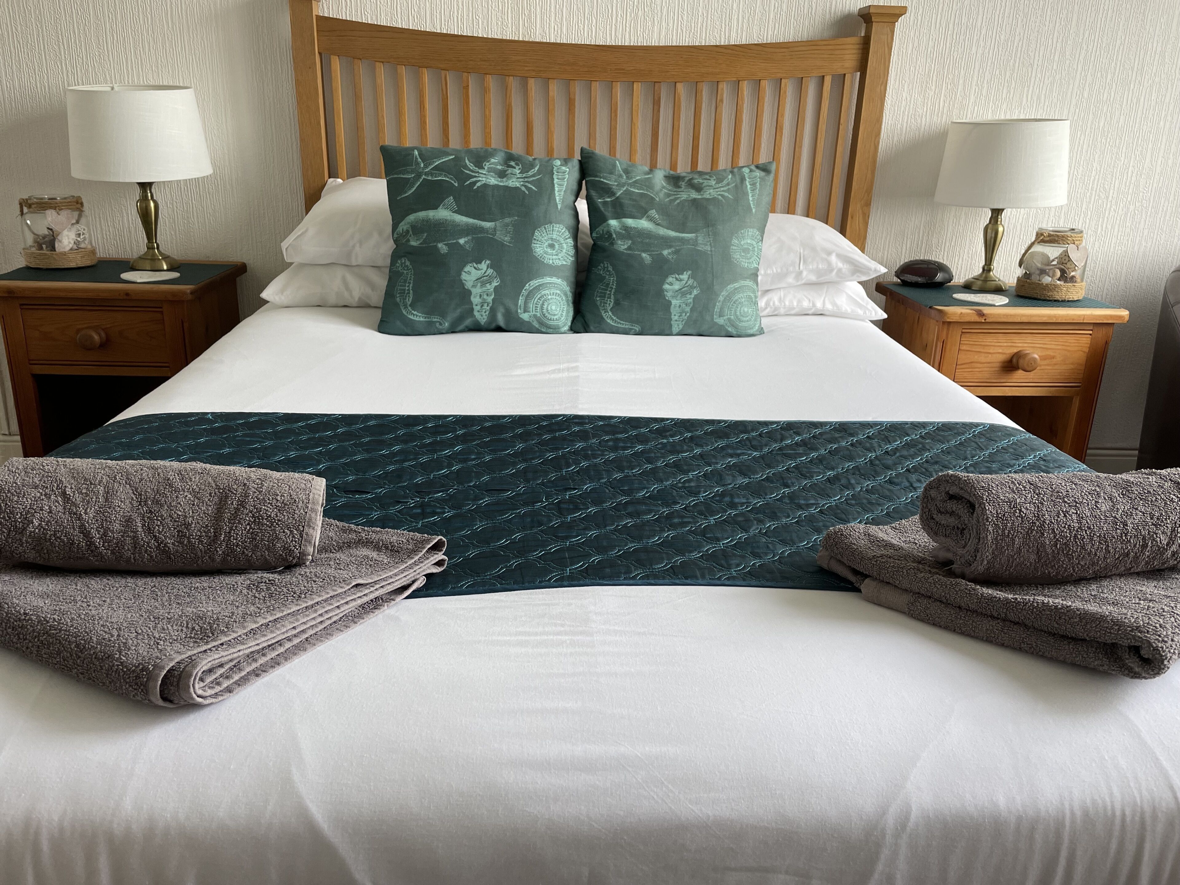 A neatly made bed with a green quilt, two pillows with a fish pattern, and two bedside tables with lamps and decorative jars.