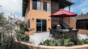 A modern two-story house with a patio area featuring a dining table and chairs, a red umbrella, and a staircase leading to the upper floor. - Scenic Waterfront Getaway Townhouse with Kayak! (Oxnard)