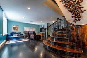 A spacious living area with a dark wooden floor, a blue sofa, a leather armchair, and a staircase with a decorative railing.