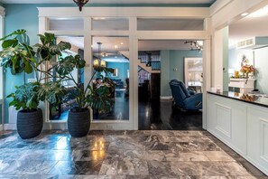 A spacious interior with a marble floor, large potted plants, a blue chair, and a kitchen area.