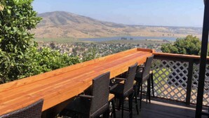 Una terraza de madera con una larga mesa y sillas, con vista a un paisaje que incluye un valle y una masa de agua.