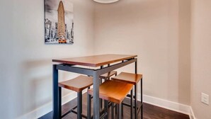 Una habitación de esquina con una mesa y dos sillas, un cuadro de un edificio, y una lámpara en el techo.