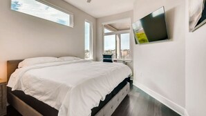 Un dormitorio moderno con una cama grande, ventilador de techo, televisor montado en la pared y grandes ventanales con vista a la ciudad.